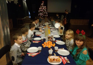 Przy uroczyście nakrytym stole siedzą uśmiechnięte dzieci w oczekiwaniu na rozpoczęcie spotkania wigilijnego.