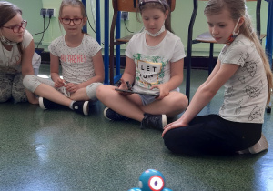 Cztery dziewczynki siedzą na podłodze i przyglądają się niebieskiemu robocikowi.