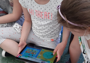 Dziewczynka siedzi skrzyżnie na podłodze. W rękach trzyma tablet i patrzy w dal. Druga dziewczynka wpatrzona jest w ekran tabletu.