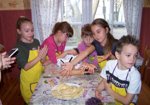 Pięcioro uczniów w fartuszkach stoi przy stole, dziewczynka wskazuje talerz własnoręcznie ulepionych pierogów.