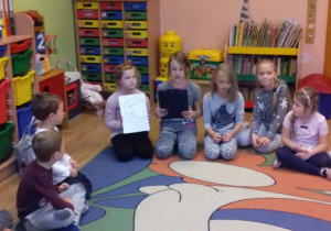 Dziewczynka czyta opowiadanie, wokół są zgromadzone młodsze dzieci - przedszkolaki.