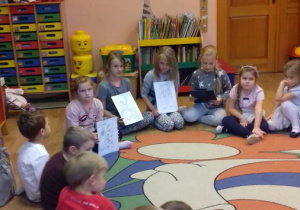 Dziewczynka czyta opowiadanie, trzy dziewczynki pokazują obrazki, wokół siedzą na dywanie przedszkolaki.