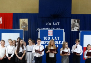Kilkoro występujących uczniów stoi na scenie, w tle widnieją zdjęcia Józefa Piłsudskiego oraz plakaty o tematyce patriotycznej związanej z wydarzeniem.