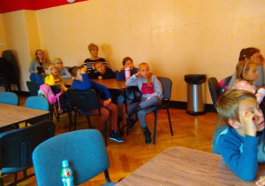Dwie dorosłe kobiety i dziesięcioro dzieci siedzi przy trzech stolikach i patrzy w prawą stronę.