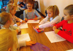 Pięć dziewczynek siedzi przy stole, w rękach trzymają kredki, a przed nimi leżą kartki do kolorowania.
