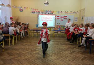 Sala lekcyjna wypełniona dziećmi, na środku sali uśmiechnięty chłopiec prezentuje swój strój w barwach narodowych.