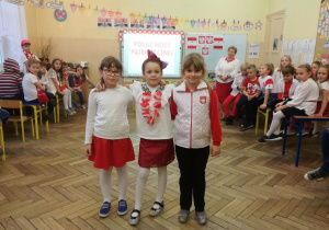Sala lekcyjna wypełniona dziećmi, środkiem sali idą trzy uśmiechnięte dziewczynki prezentując swoje stroje w barwach biało - czerwonych