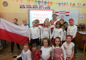 Sala lekcyjna, przed tablicą grupa uśmiechniętych uczniów w biało - czerwonych strojach. Chłopiec z lewej strony prezentuje flagę Polski.