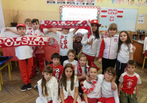 Sala lekcyjna, przed tablicą grupa uśmiechniętych uczniów prezentuje biało - czerwone stroje ,szaliki i inne akcesoria w barwach narodowych.