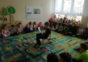 Sala przedszkolna, pośrodku sali otoczony dziećmi siedzi czytający wiersz chłopiec.