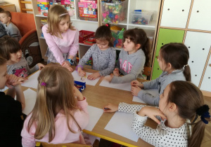 Sala przedszkolna, przy stoliku siedzą dzieci, które wspólnie wykonują prace pastyczne.