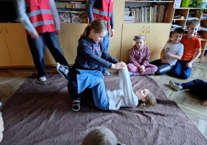 Sala lekcyjna, dziewczynki prezentują udzielanie pierwszej pomocy przedmedycznej podczas omdlenia.