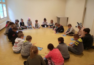 Sala muzyczna, siedząca w kręgu grupa uśmiechniętych uczniów pod okiem instruktorki gra na dzwonkach.