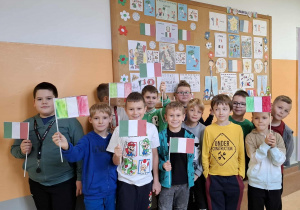 Korytarz szkolny, uśmiechnięte dziewczynki prezentują samodzielnie wykonane flagi Włoch oraz tablicę z symbolami Italii.