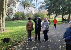 Jesienny park, dzieci obserwują ptaki i wiewiórki z wykorzystaniem lornetki.