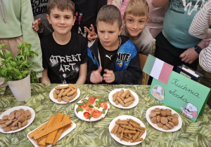 Sala lekcyjna, uśmiechnięci uczniowie zgromadzeni przy stole pełnym włoskich przysmaków.