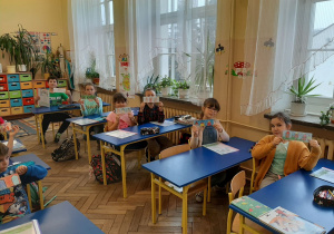 Sala lekcyjna, uśmiechnięci siedzący w ławkach uczniowie prezentują wykonane samodzielnie kolorowe zakładki do książek, nawiązujące do przeczytanej lektury "Dzieci z Bullerbyn".